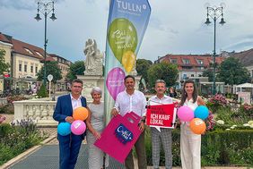Fünf Personen halten bunte Luftballons und Einkaufstaschen vor einer Fahne mit der Aufschrift "TULLN" in Tulln.